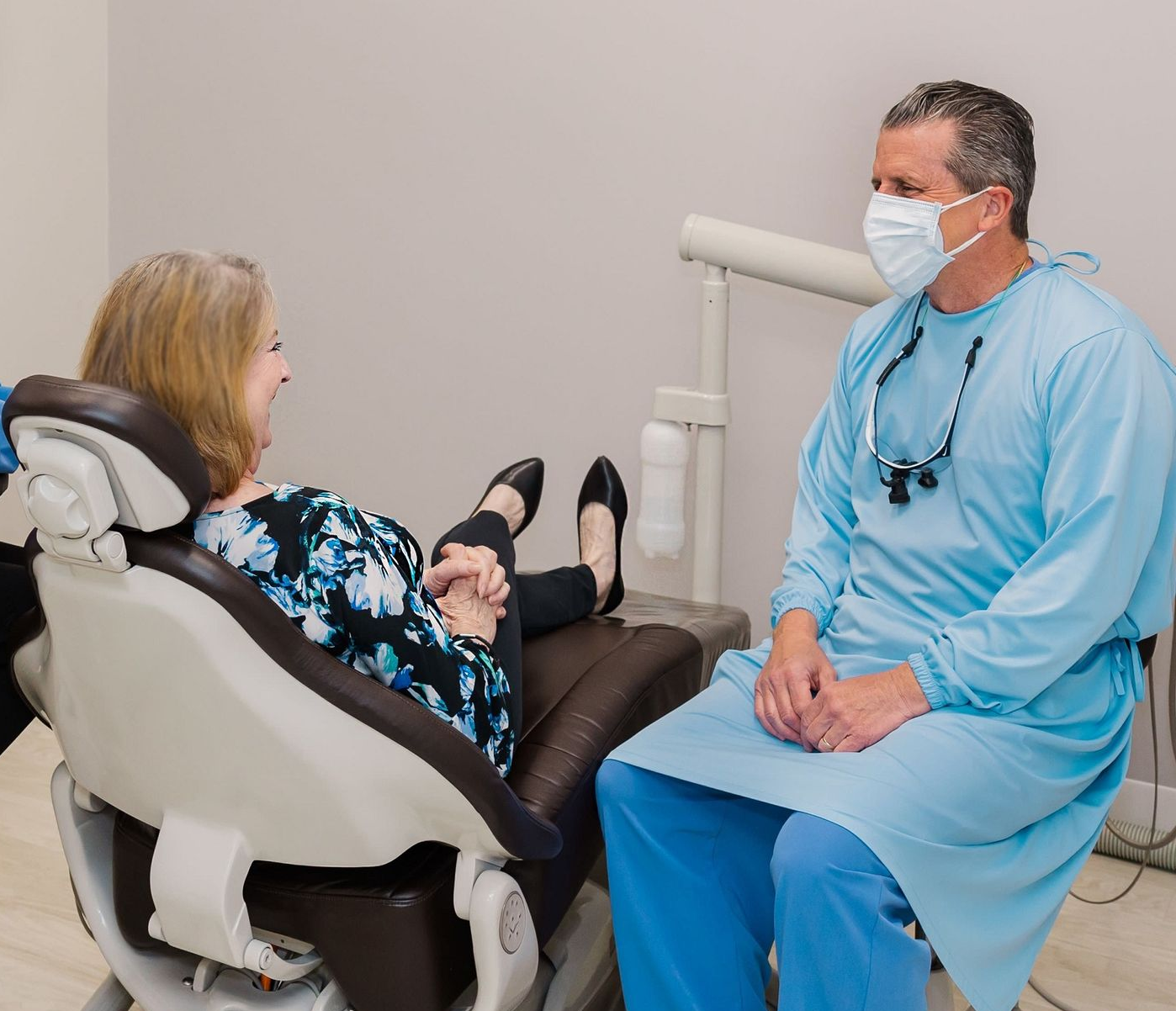 Dental exam with patient in chair, dentist in blue scrubs and masks talking in a clinic room.