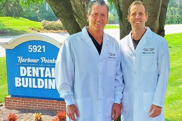 Two dentists in white coats standing beside a blue dental building sign outdoors