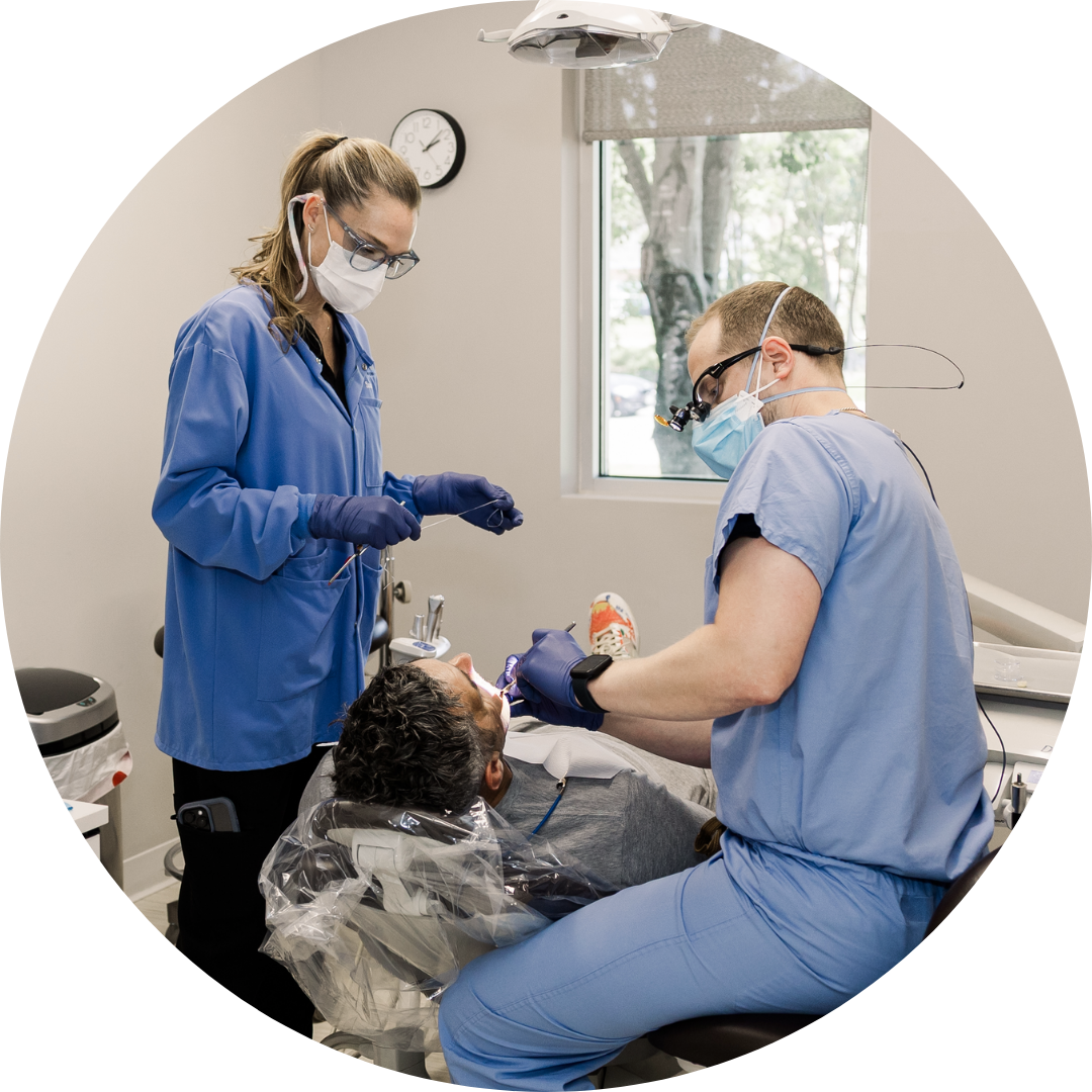 Dentist and assistant treating a patient in a dental chair, wearing blue scrubs and masks.