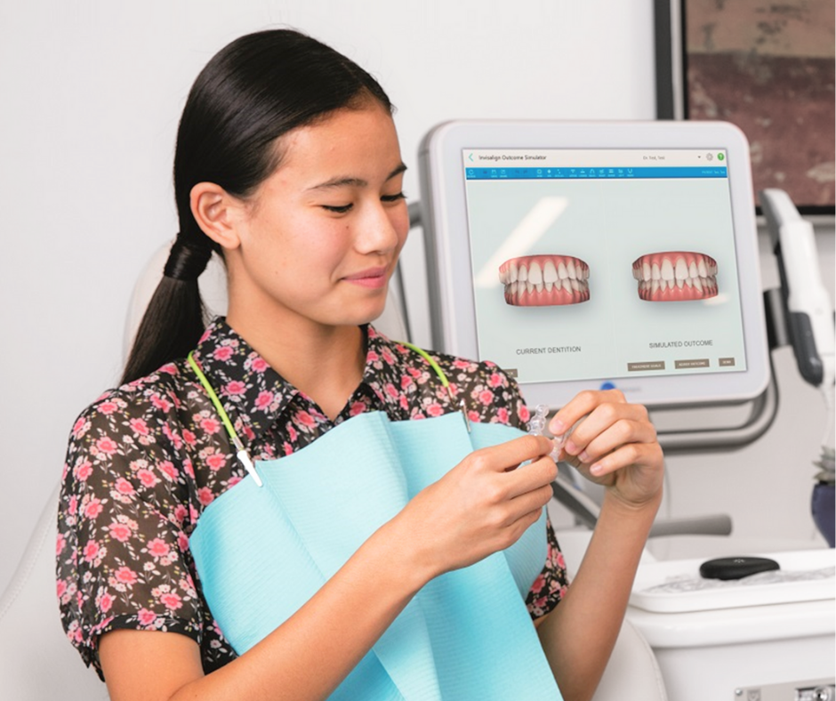 Woman in a dental office holding aligners, with a computer showing tooth models in the background.