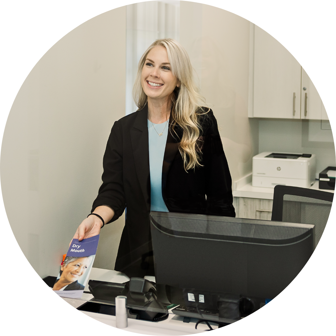 Smiling receptionist at a front desk, holding a brochure beside a computer in a bright office.