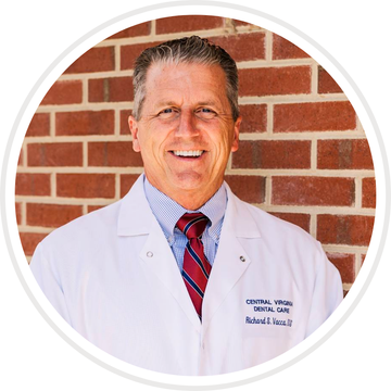 Doctor in white coat and tie standing in front of a brick wall, smiling.