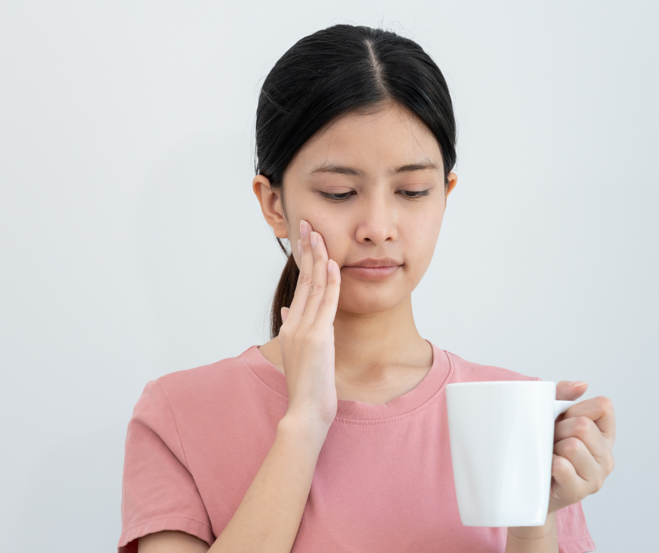 Woman holding a white mug and touching her cheek, looking uncomfortable against a plain light background