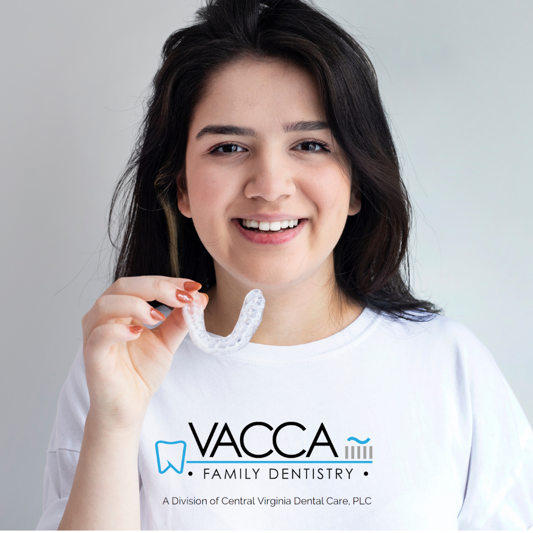 Woman smiling in a white Vacca Family Dentistry shirt, holding a dental aligner against a light background