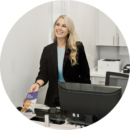 Smiling woman behind a desk in a bright office, holding a brochure beside a computer monitor