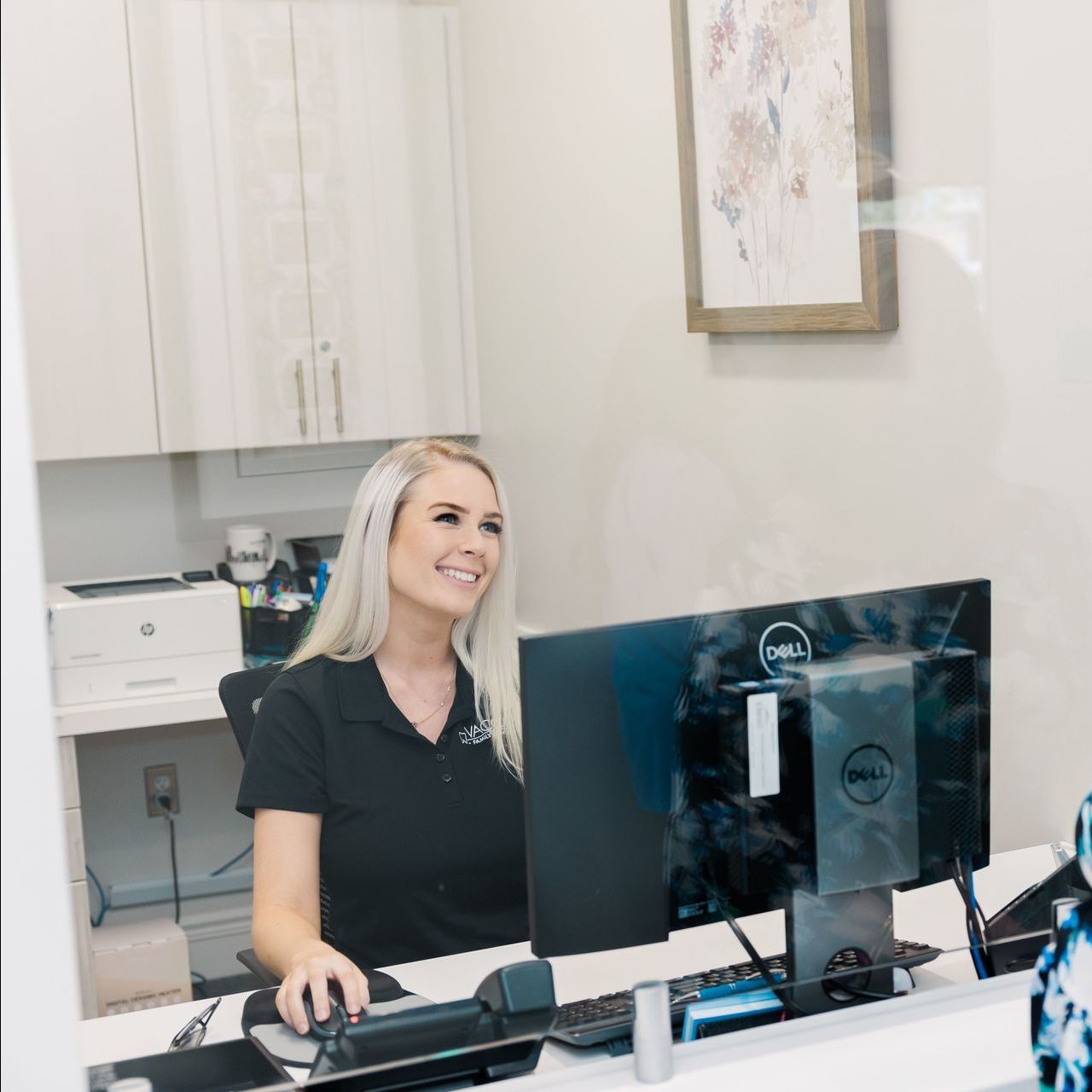Woman smiling at a desk in a bright office, working at a computer and phone setup.