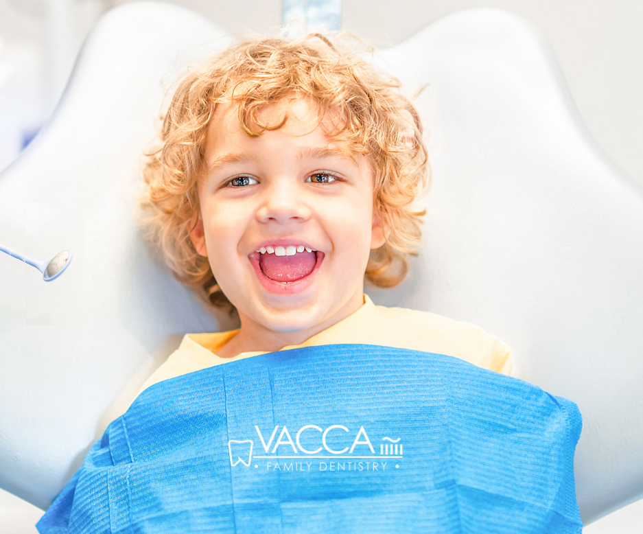A smiling young child with curly blonde hair sits in a dentist's chair, wearing a blue bib.