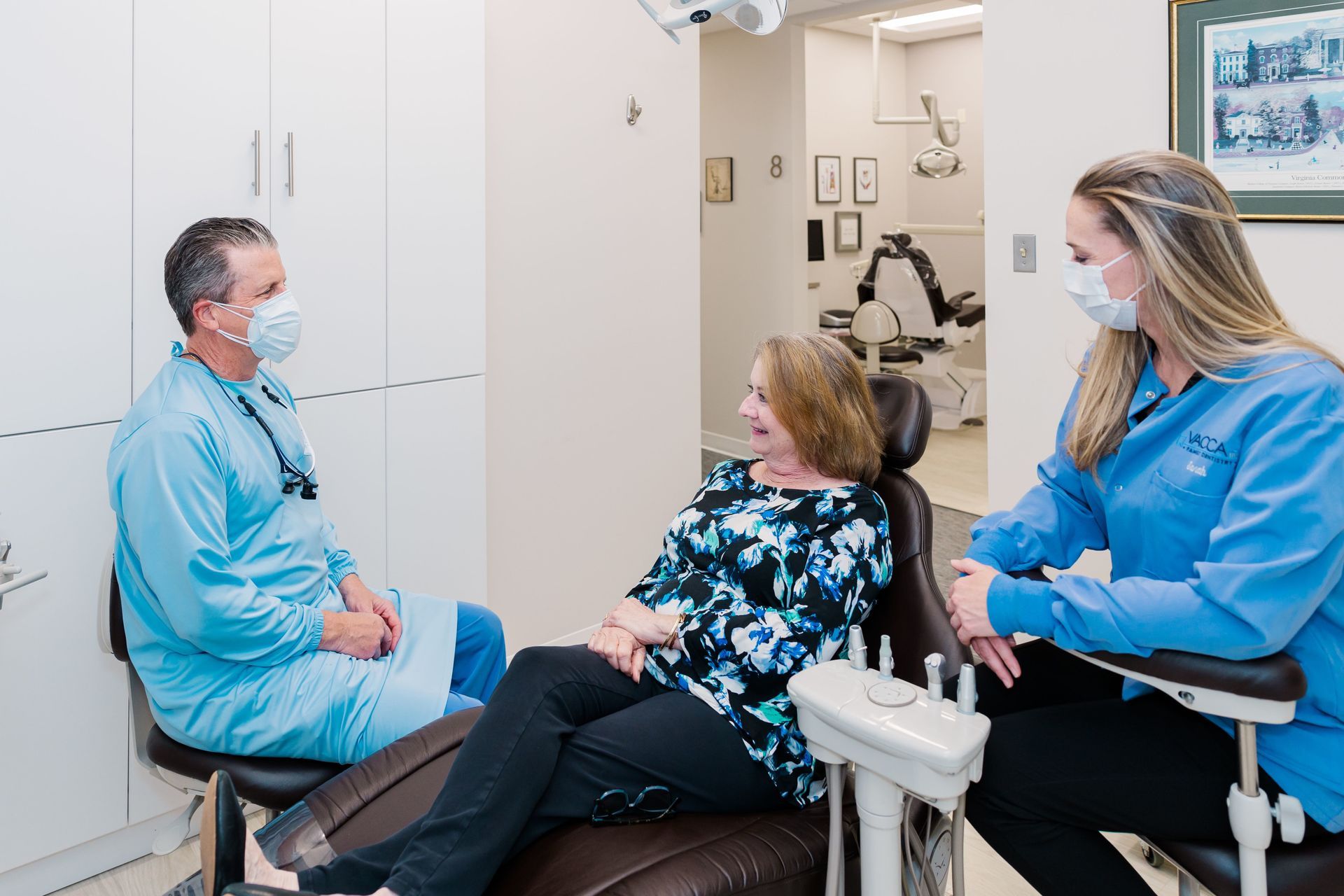 Two clinicians talking with a seated patient in a dental exam room, all wearing masks.