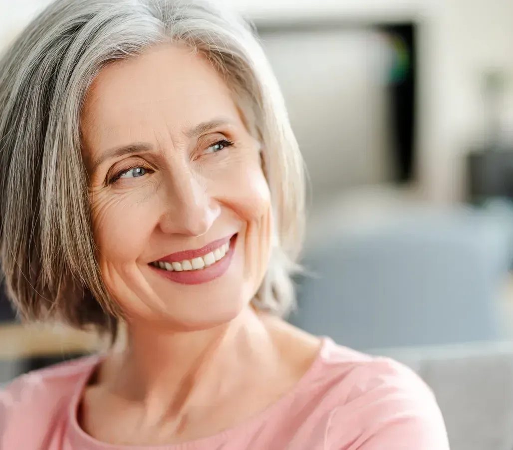 Smiling woman with short gray hair in a pink shirt, seated indoors against a blurred background
