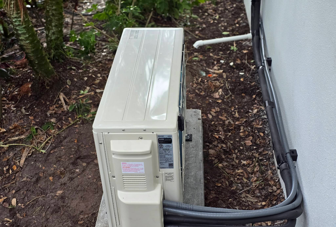 Outdoor air conditioner unit beside a house wall, with a fan grill and beige casing.
