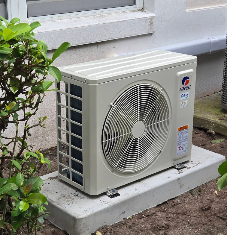 Outdoor air conditioner unit beside a house wall, with a fan grill and beige casing.