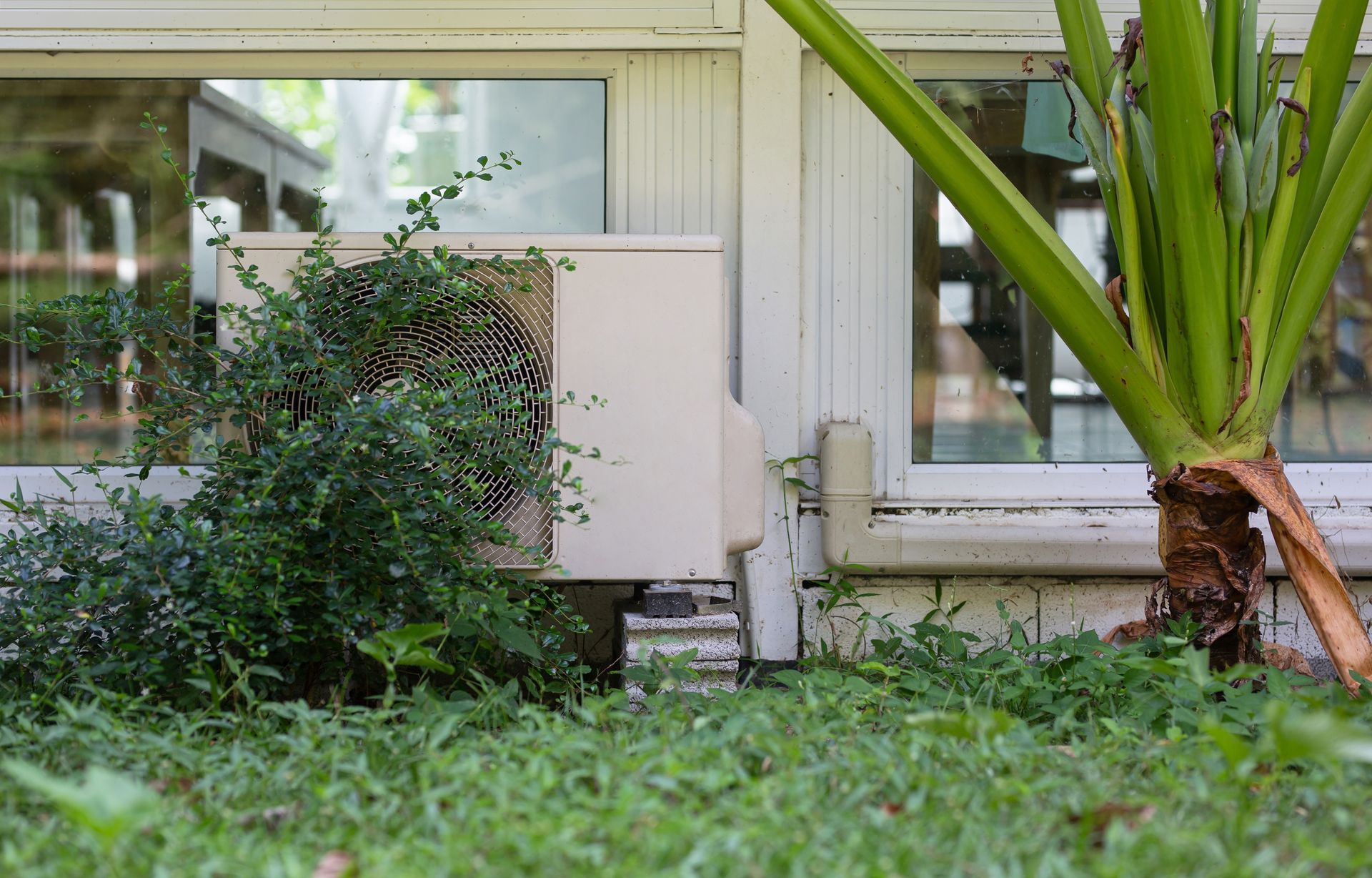 Outdoor air conditioner unit beside a house wall, with a fan grill and beige casing.