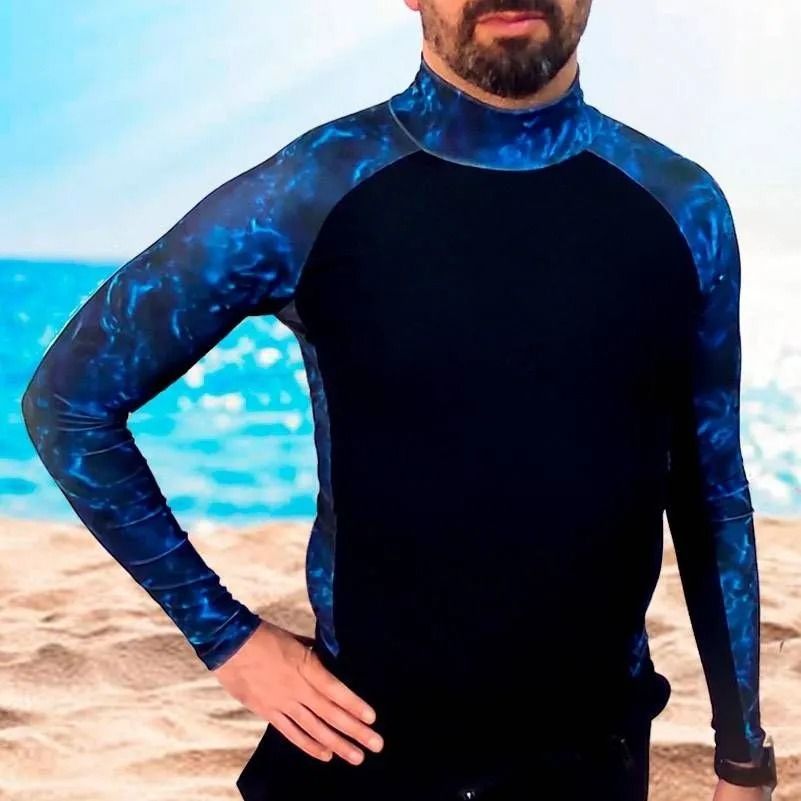 Hombre con una camiseta de licra azul y negra, de pie en una playa con el océano detrás