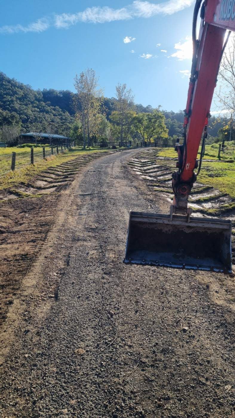 An excavator working on a gravel road with a green hill and trees in the background. Blue sky.