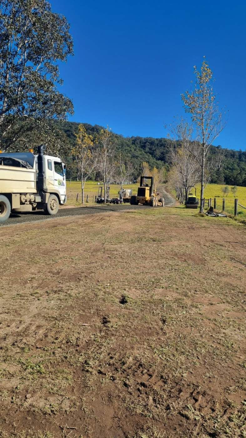 Truck and yellow bulldozer on a dirt path near a grassy field, trees, and a mountain under a blue sky.