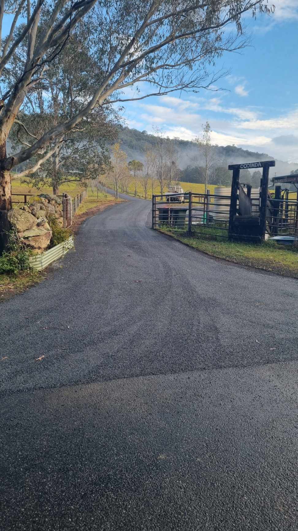 Freshly paved farm driveway leading past a gate toward rolling hills and trees under a blue sky.— Hunter Valley Bitumen In Gresford, NSW