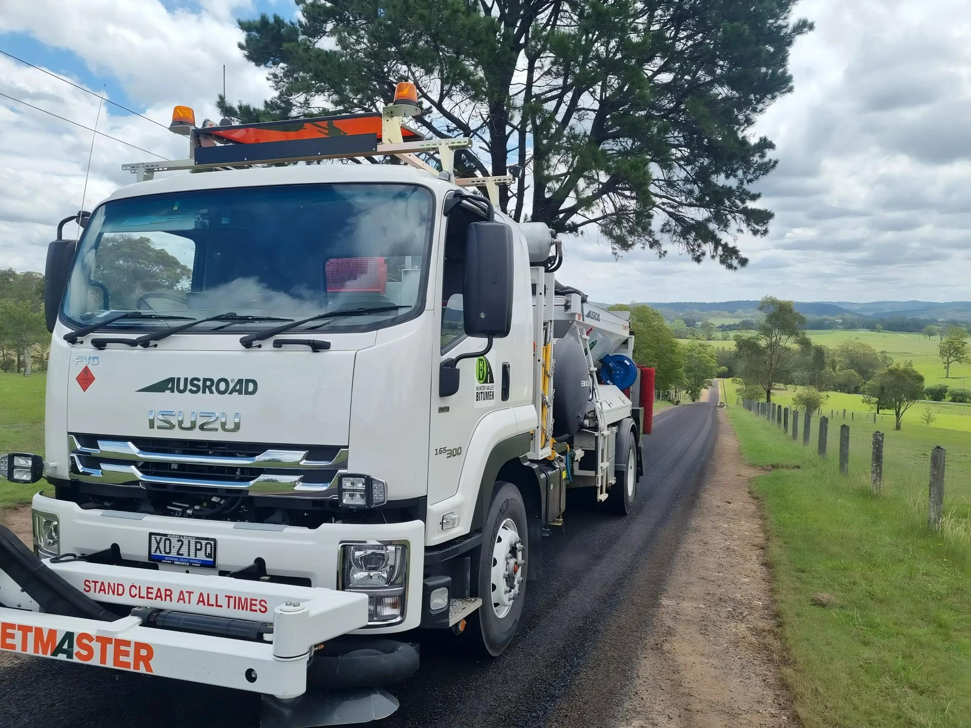 White Ausroad Isuzu truck on a rural road with green fields and hills in the background. — Hunter Valley Bitumen In Maitland, NSW