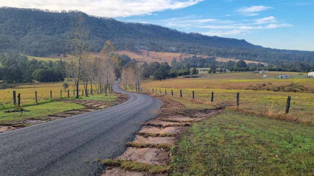 Gravel road curves through a field towards a tree-lined hill under a blue sky.