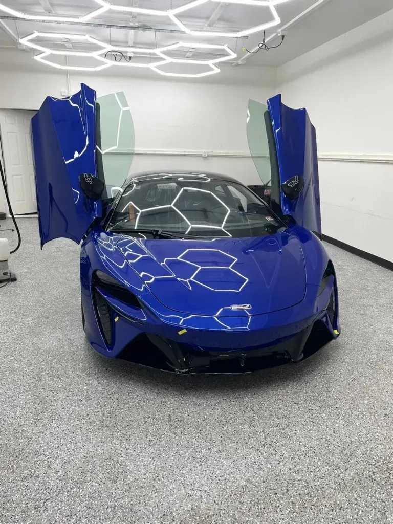 Blue sports car in a white showroom with butterfly doors open, viewed from the front.