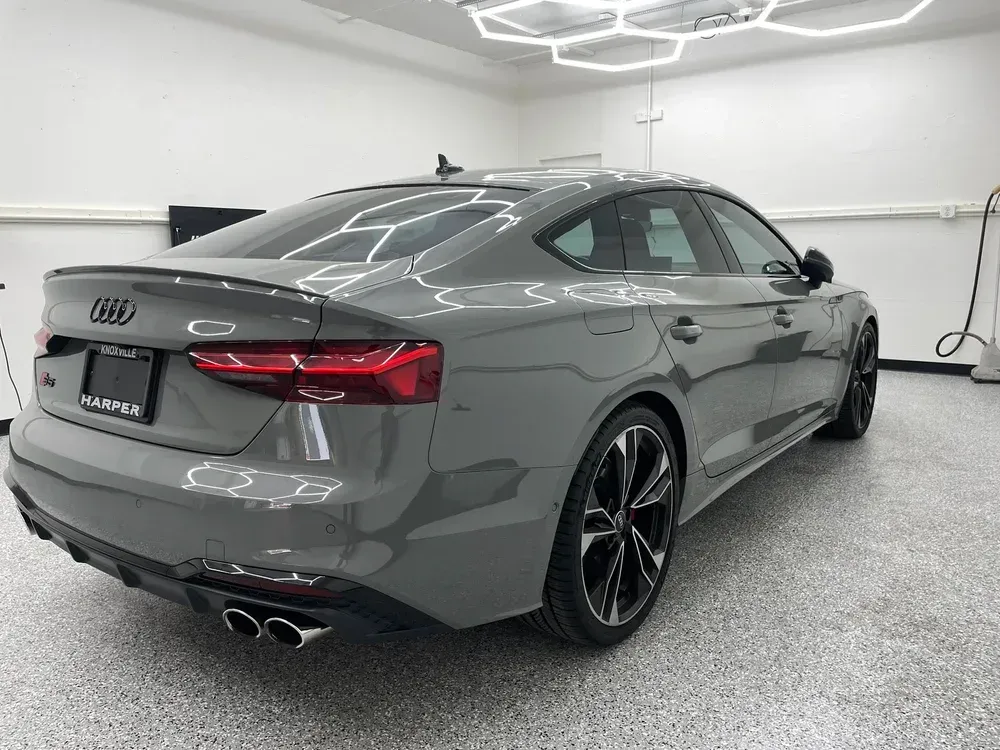 Gray Audi sedan in a white garage, rear three-quarter view with black rims and dual exhausts.