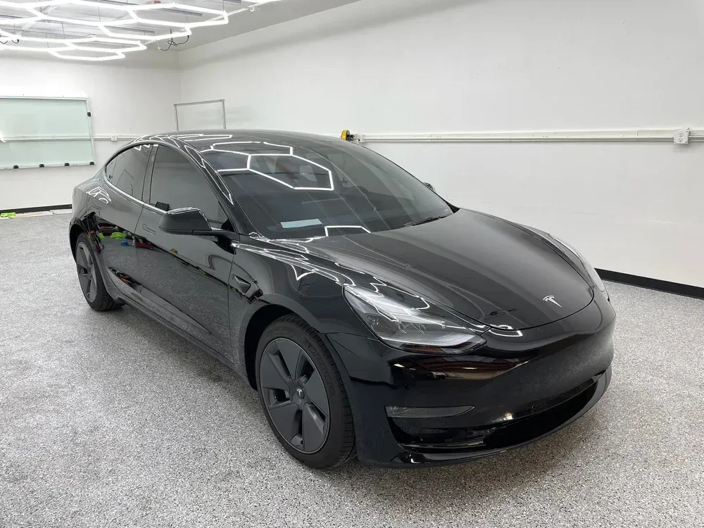 Black Tesla sedan in a bright white garage, viewed from the front-left side.