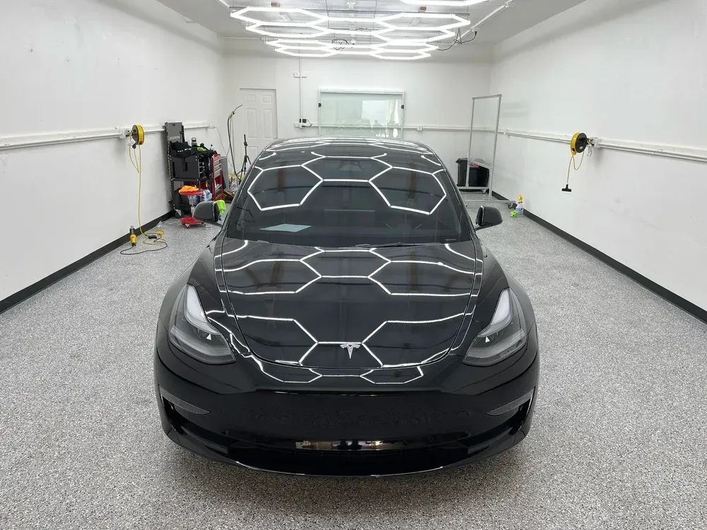 Black car in a white detailing garage with hexagonal ceiling lights reflected on the hood.