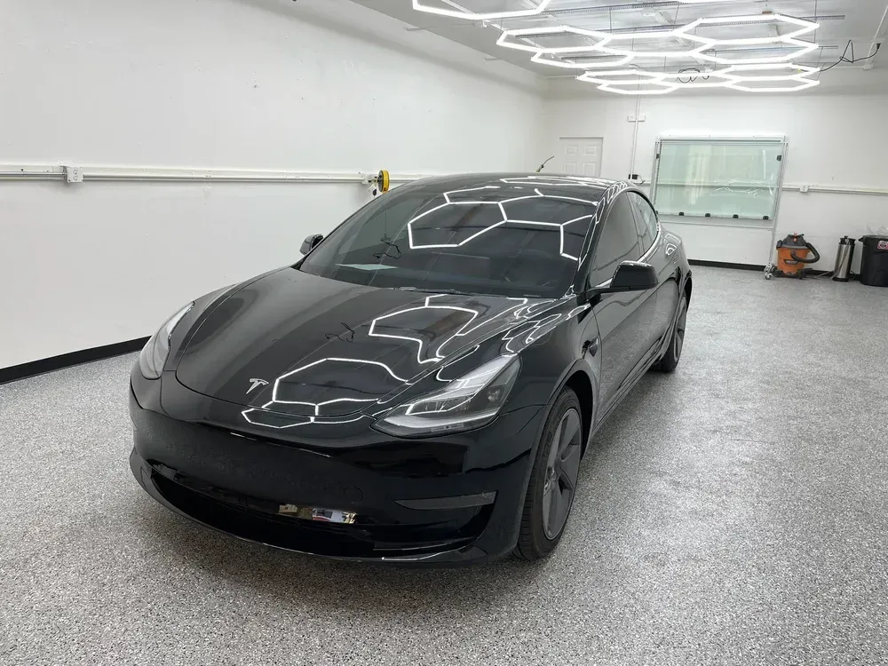 Black Tesla sedan in a garage with bright overhead lights reflecting on the hood.