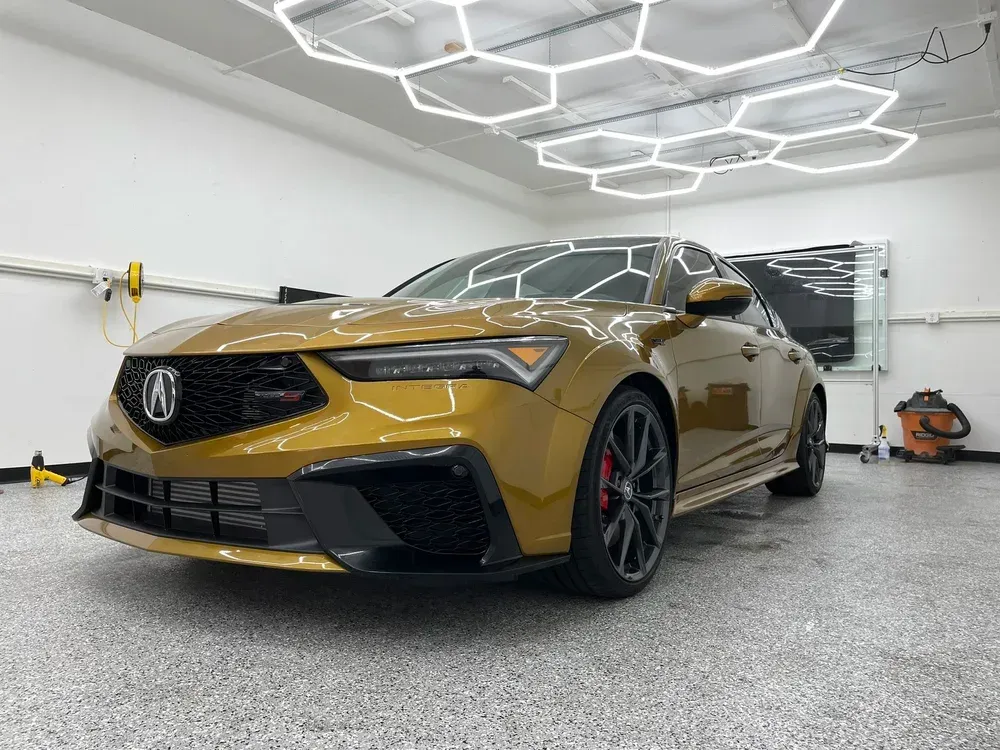 Gold Acura sedan in a modern garage, front three-quarter view under geometric ceiling lights.