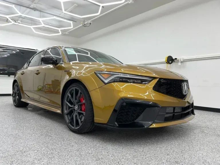 Gold Acura sedan in a garage, front three-quarter view, with black wheels and red brake calipers.
