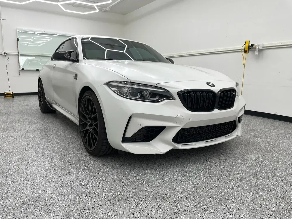 White BMW coupe in a clean garage, front three-quarter view, with black rims and a speckled floor.
