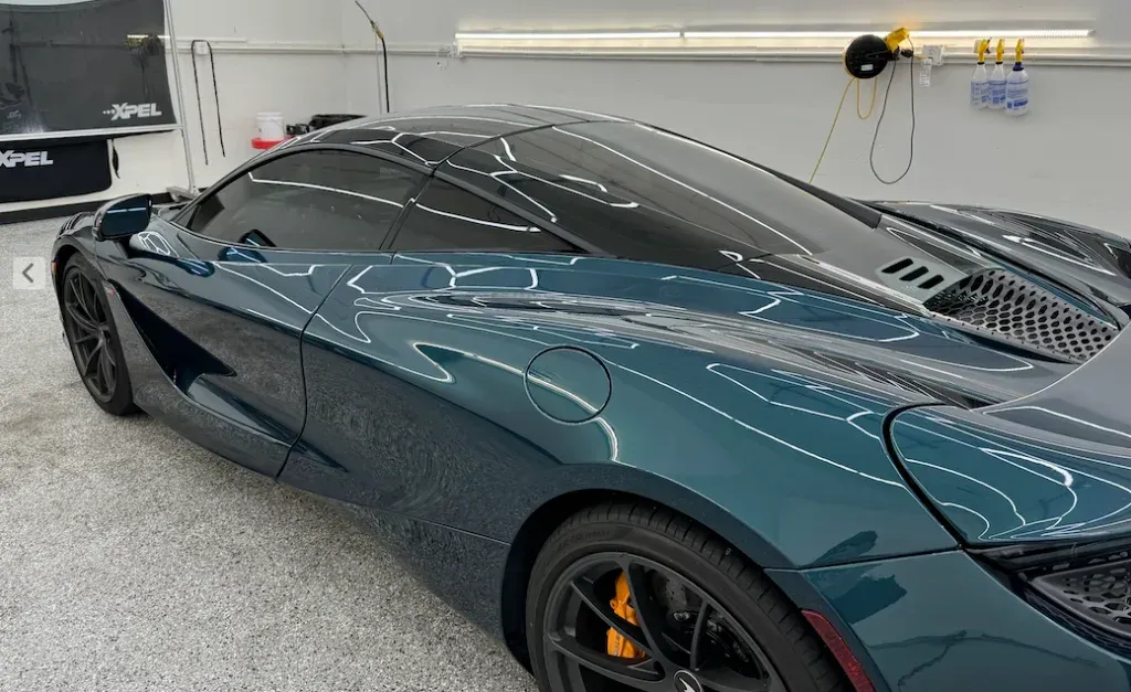Sleek dark teal sports car in a garage, seen from the side under bright overhead lights.