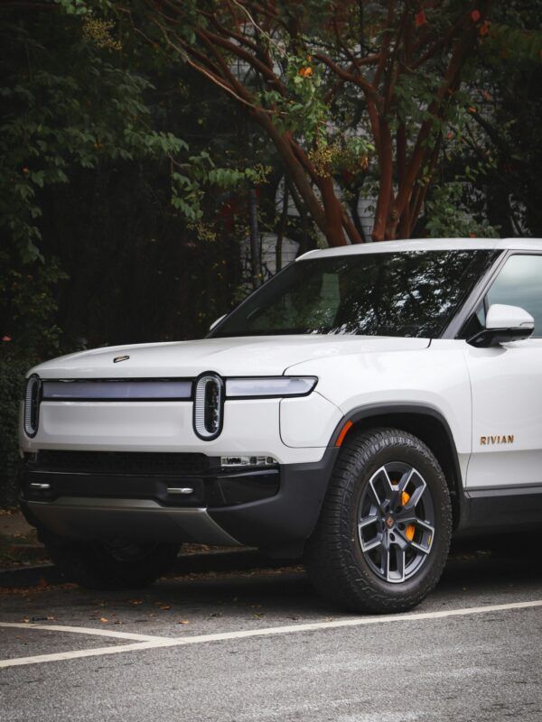 White Rivian electric SUV parked on a wet street beside trees, front three-quarter view