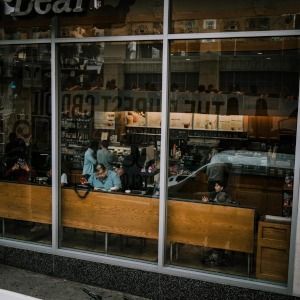 Café window with seated patrons inside, wooden bench seating, and reflections of the street outside.