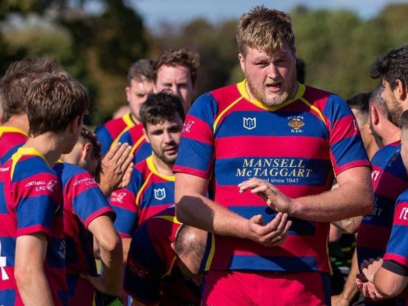 A man in a mansell mccaggart shirt is talking to a group of rugby players.