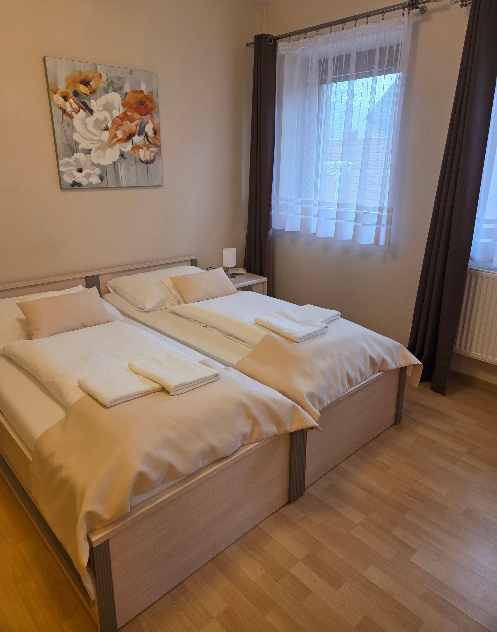 Dve úhľadne ustlané jednolôžka v béžovej spálni s oknom a kvetinovou nástennou maľbou.