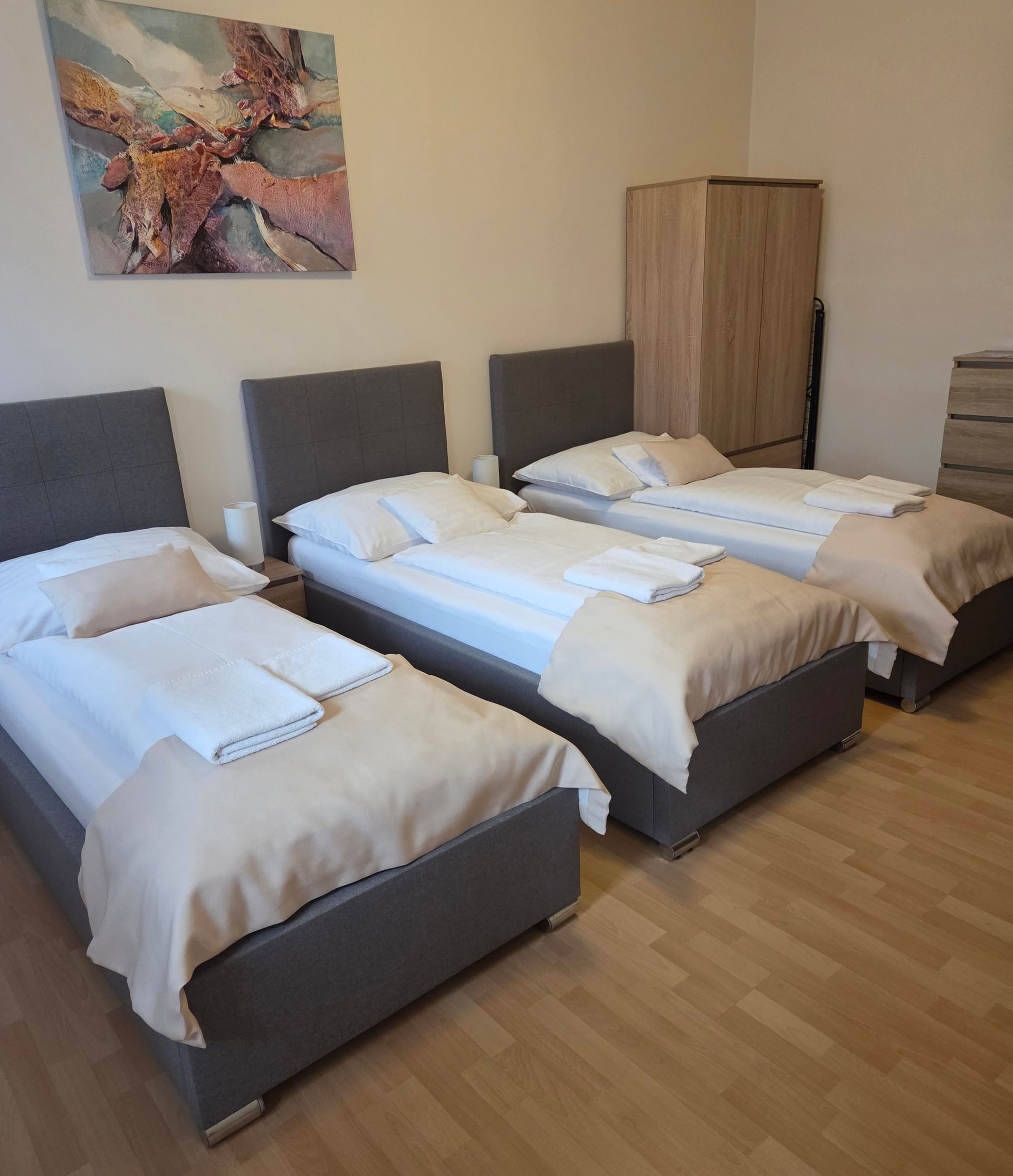 Tri samostatné postele v upratanej spálni so sivými čelami postelí, bielou posteľnou bielizňou a drevenou skriňou
