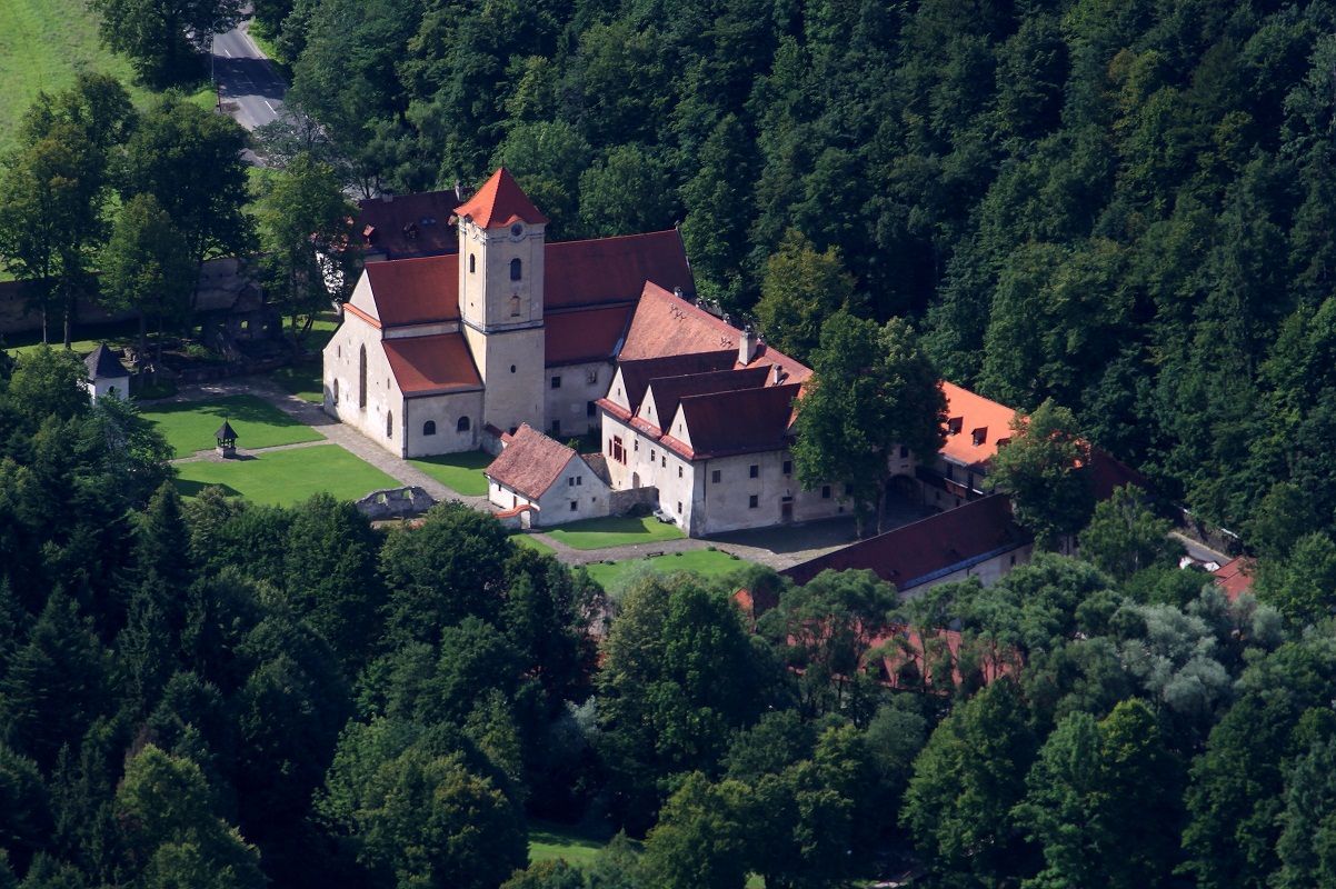 Autor: Kamil Gwóźdź – Vlastní dílo, CC BY-SA 3.0, https://commons.wikimedia.org/w/index.php?curid=17887764 Letecký pohľad na biely hrad s červenými strechami obklopený hustým zeleným lesom