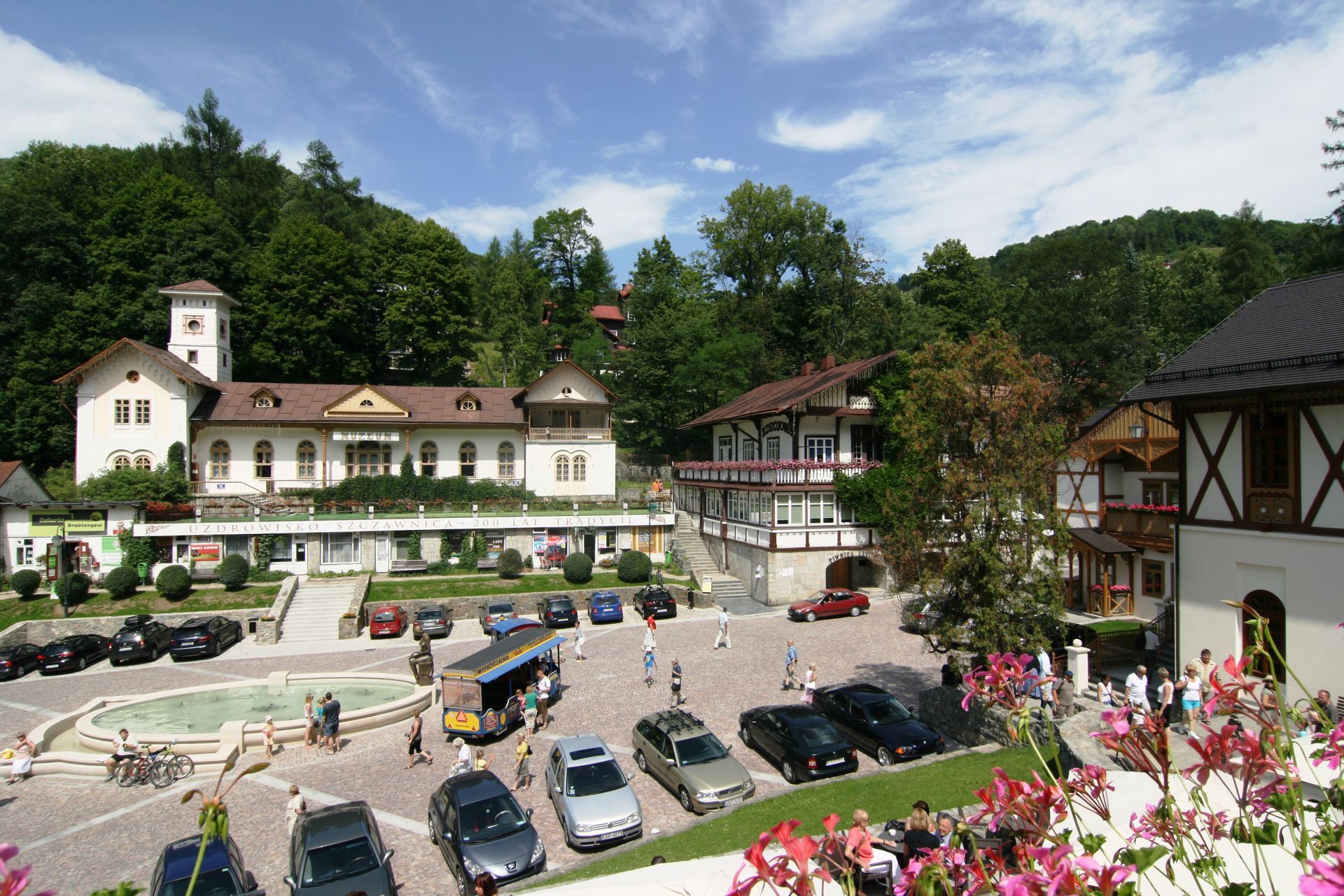 Zdroj: https://pl.wikipedia.org/wiki/Szczawnica#/media/Plik:PL_Szczawnica_Plac_Dietla_1.JPG Námestie horskej dediny so zaparkovanými autami, historickými budovami a kvetináčmi pod slnečnou oblohou