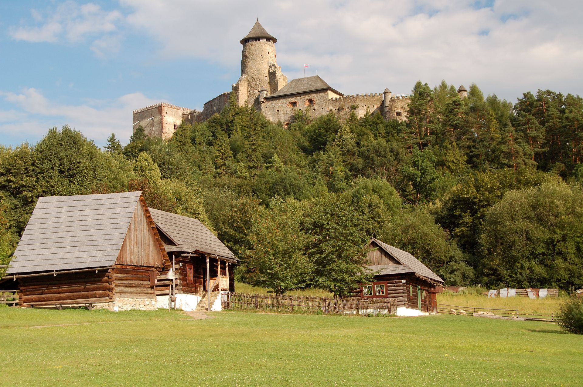 Autor: Miro Svorc – VY-SA 3.lastní dílo, CC B0, Zdroj: https://commons.wikimedia.org/w/index.php?curid=28126562 Drevené chatky na zelenom poli pod kamenným hradom na zalesnenom kopci