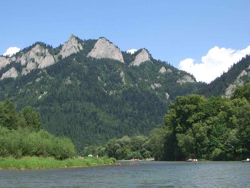 Horské štíty nad pokojnou riekou, so sviežimi zelenými stromami a modrou oblohou s oblakmi.
