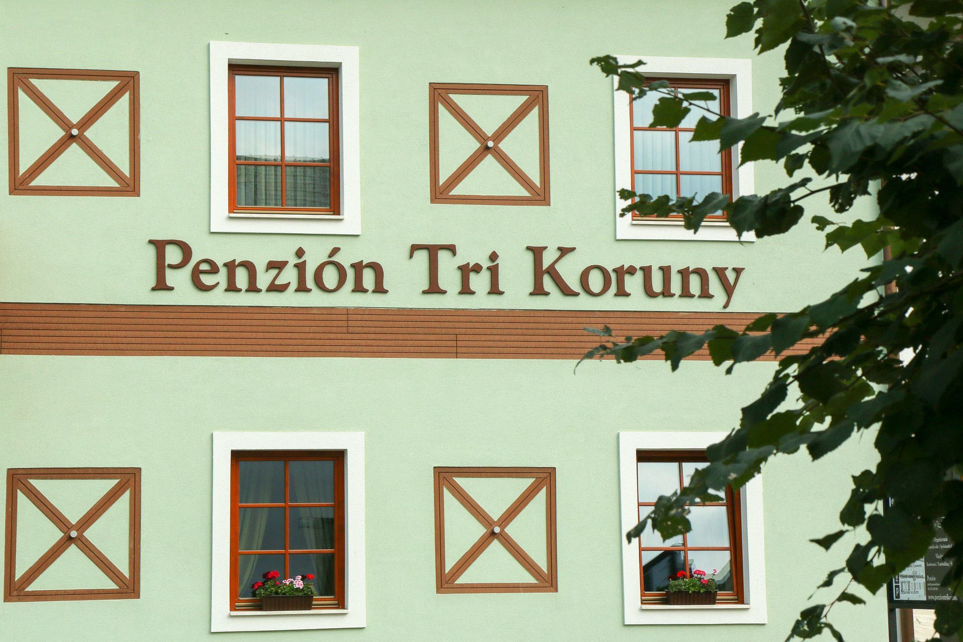 Bledozelená fasáda budovy s nápisom „Penzion Tri Koruny“ a oknami orámovanými hnedým lemom, čiastočne zakrytými listami