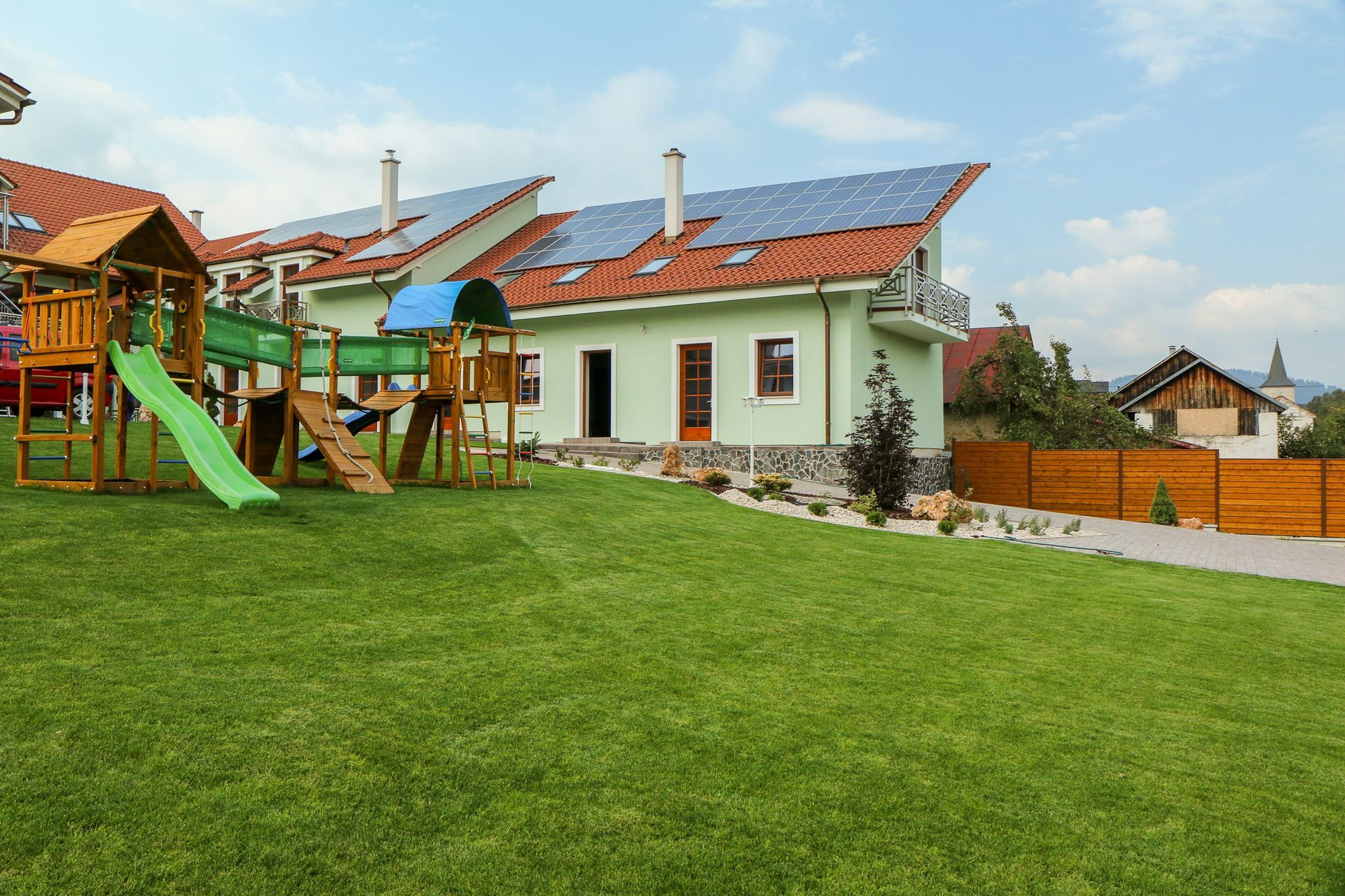Záhrada so zeleným trávnikom, ihriskom a bielym domom s červenou strechou pod modrou oblohou