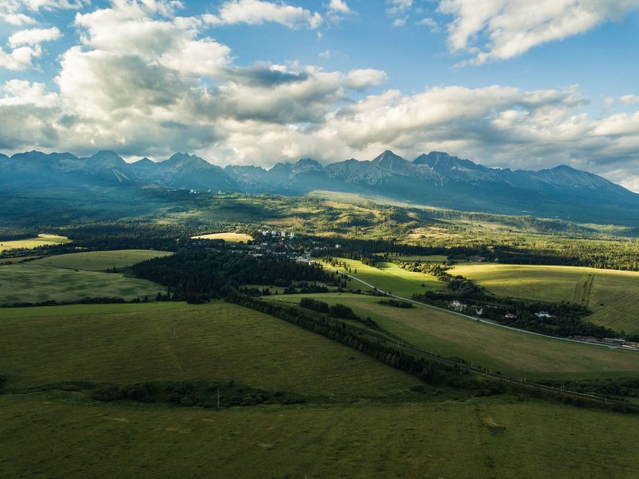 Zelené polia v údolí pod horami a oblakmi, so slnečným žiarením v krajine