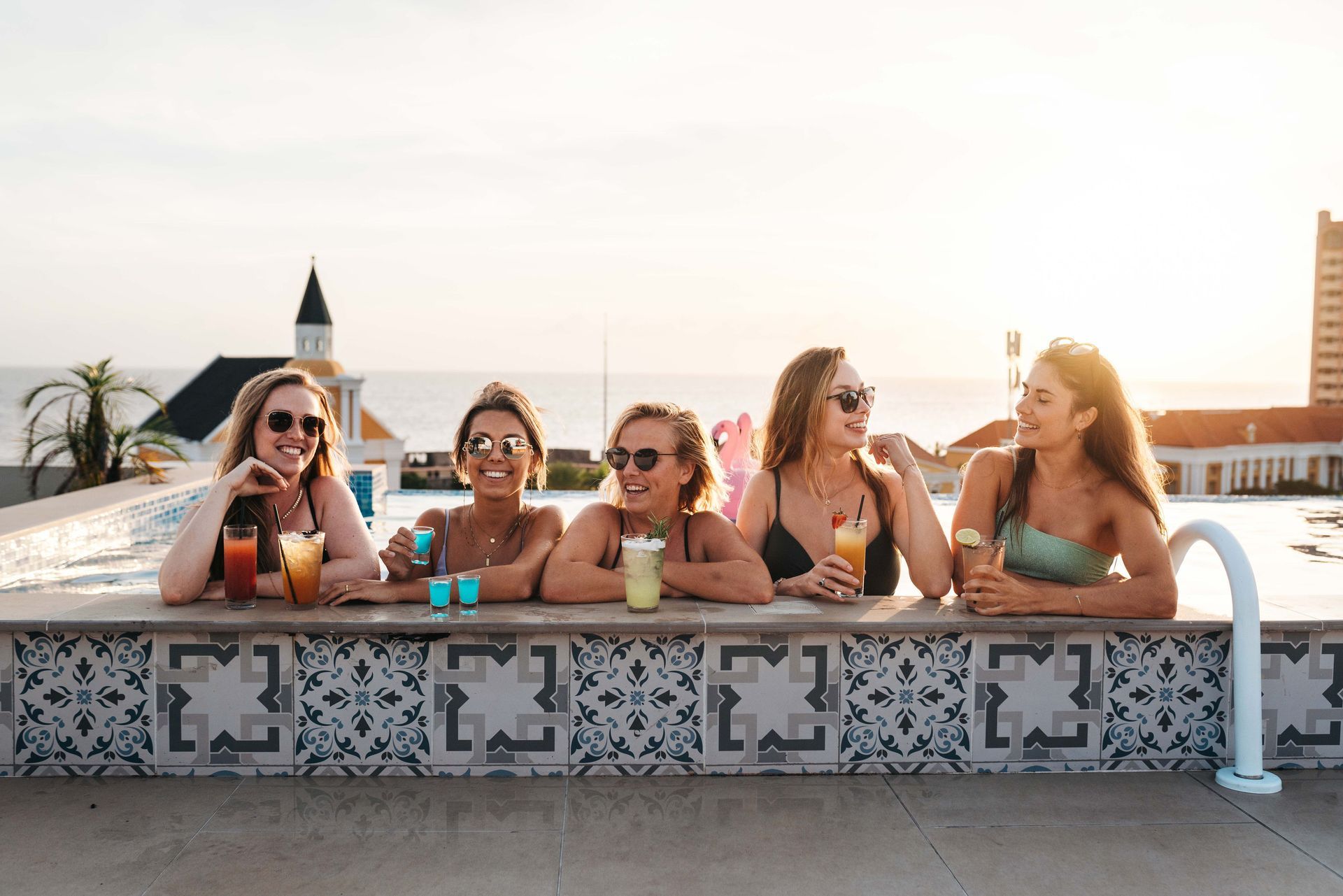 A group of women are sitting on the edge of a swimming pool drinking cocktails.