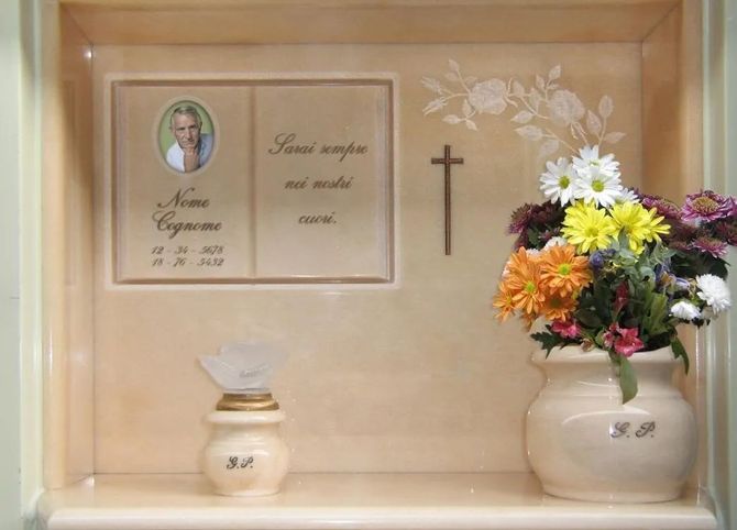 Nicchia commemorativa con targa incorniciata, croce, fiori e urna su una mensola color crema.