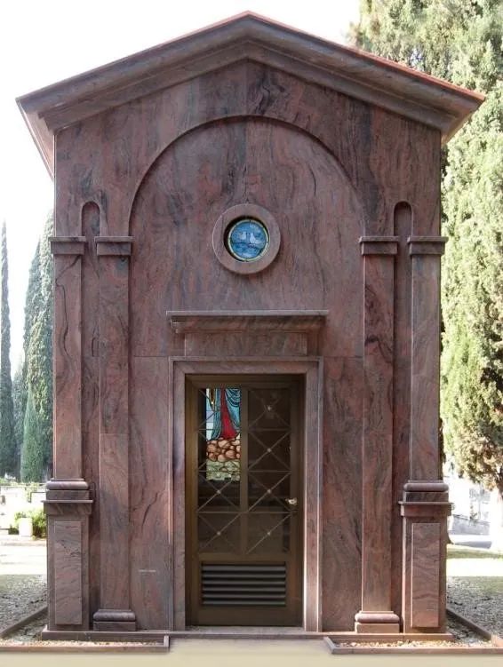 Piccolo mausoleo rosso-marrone con portale ad arco e finestra circolare tra gli alberi del cimitero