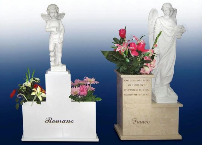 Due statue di angeli su piedistalli commemorativi con fiori rosa, uno bianco e uno beige, su uno sfondo con sfumatura blu.