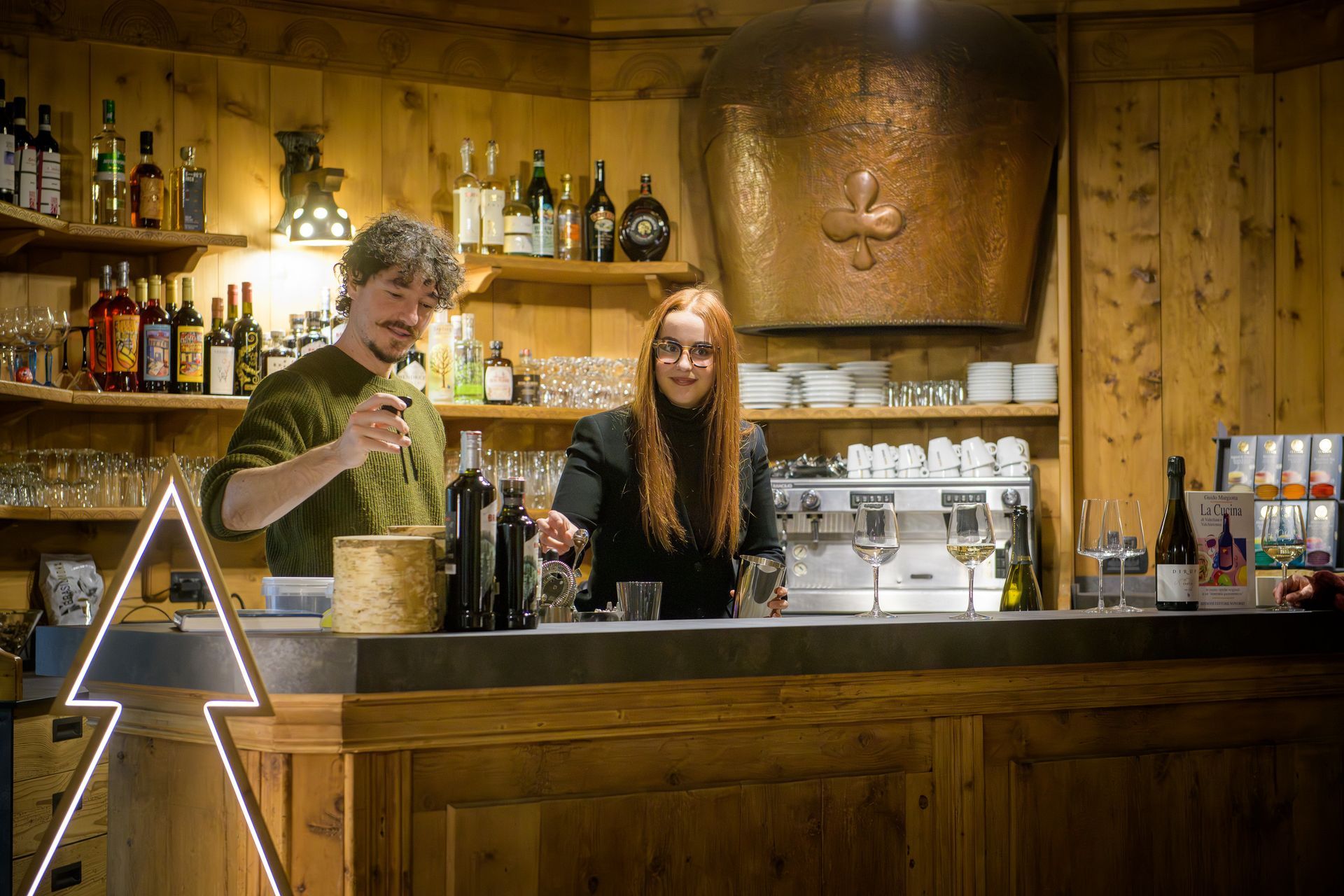 Due baristi dietro un bancone di legno in un accogliente pub, con bottiglie e bicchieri sugli scaffali alle loro spalle.