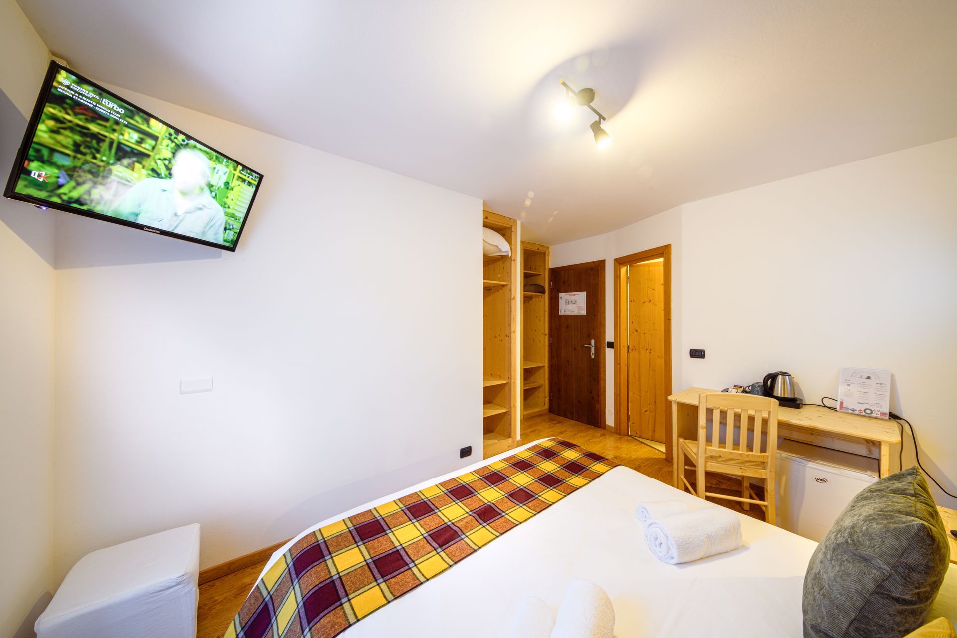 Piccola camera da letto con letto, TV a parete, armadio e scrivania con bollitore vicino alla finestra.