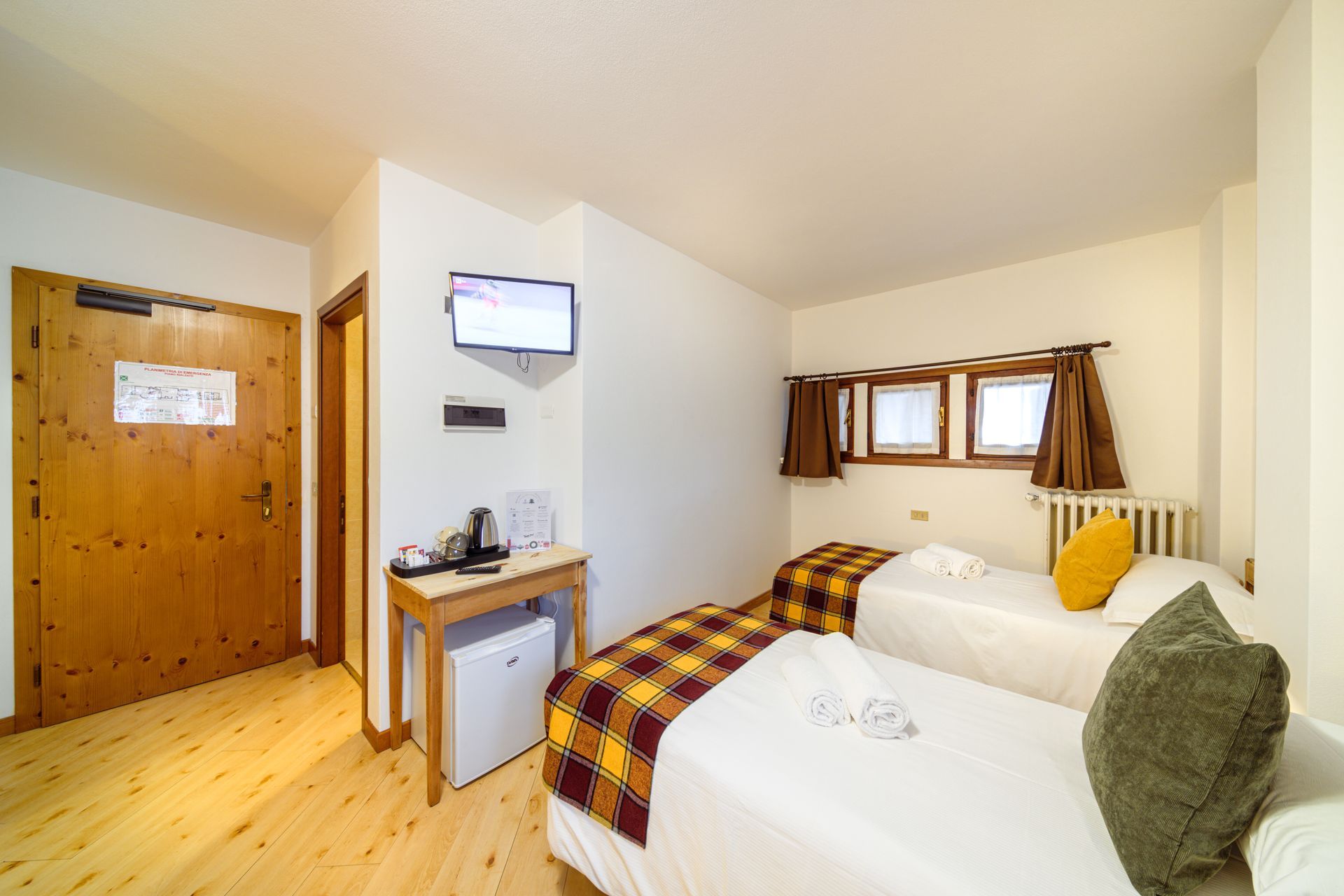 Piccola camera da letto con due letti, porta con pannelli in legno, finestra e televisore a parete.
